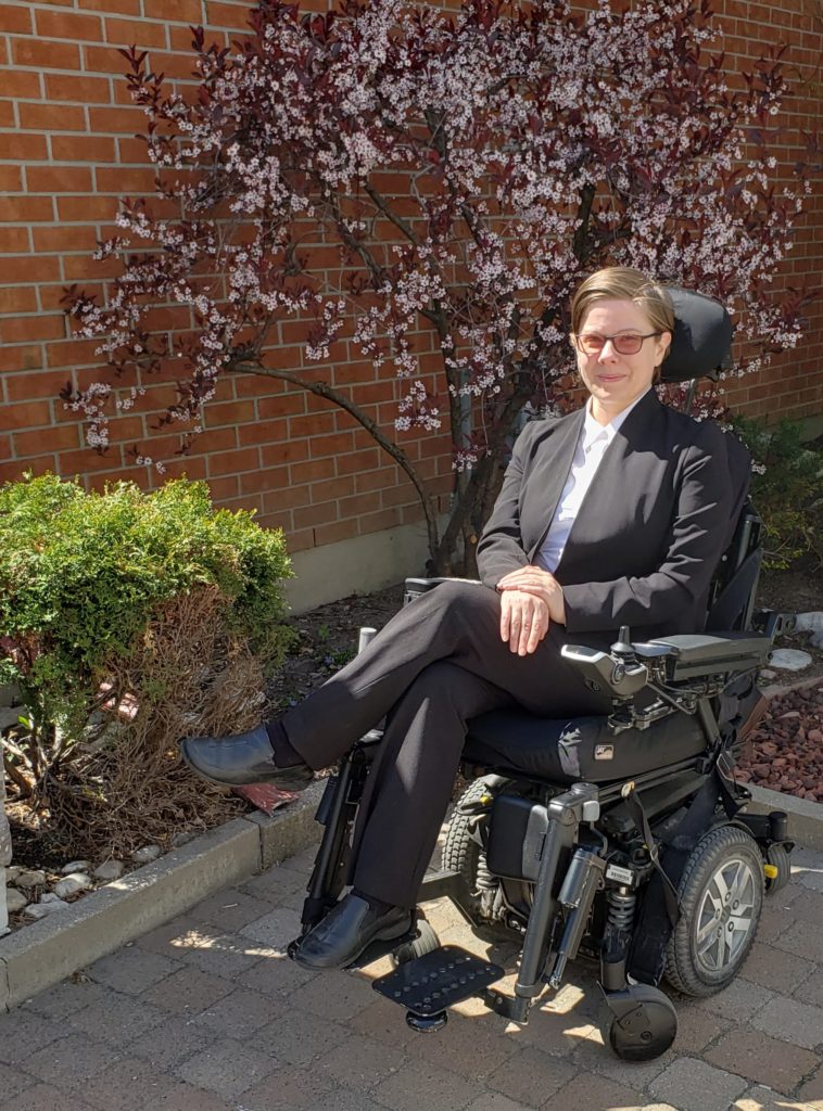 Dr. Melanie Coughlin sits comfortably in a power wheelchair with one leg crossed. She is wearing a black suit and white dress shirt. She has tinted glass and short hair. There is a flowering tree behind her and sunlight lights the photo.