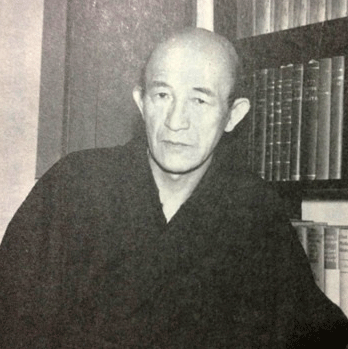 A middle-aged Japanese man sits in traditional dress next to a book case, looking calmly into the camera. 